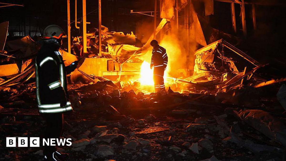 Emergency workers stand in front of a blaze at night. reflective strips on their coats stand out in the shadows and light from the fire.
