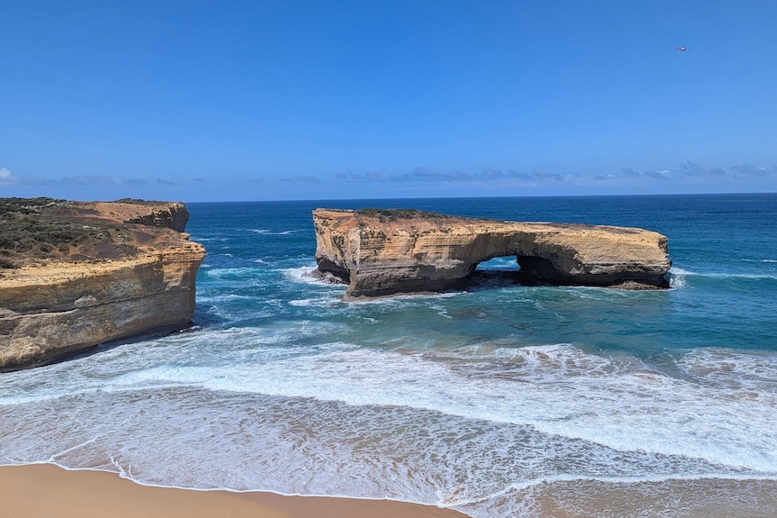 A wide-angle shot of an offshore rock formation in an aquamarine sea under clear, blue sky. 