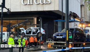 Members of a vehicle recovery team wearing yellow and orange hi-vis jackets. They are standing near the scene of the crash where a white Honda Jazz is being taken away on the bed of a crane truck. The blue BMW is visible on the right, also being removed from the scene.