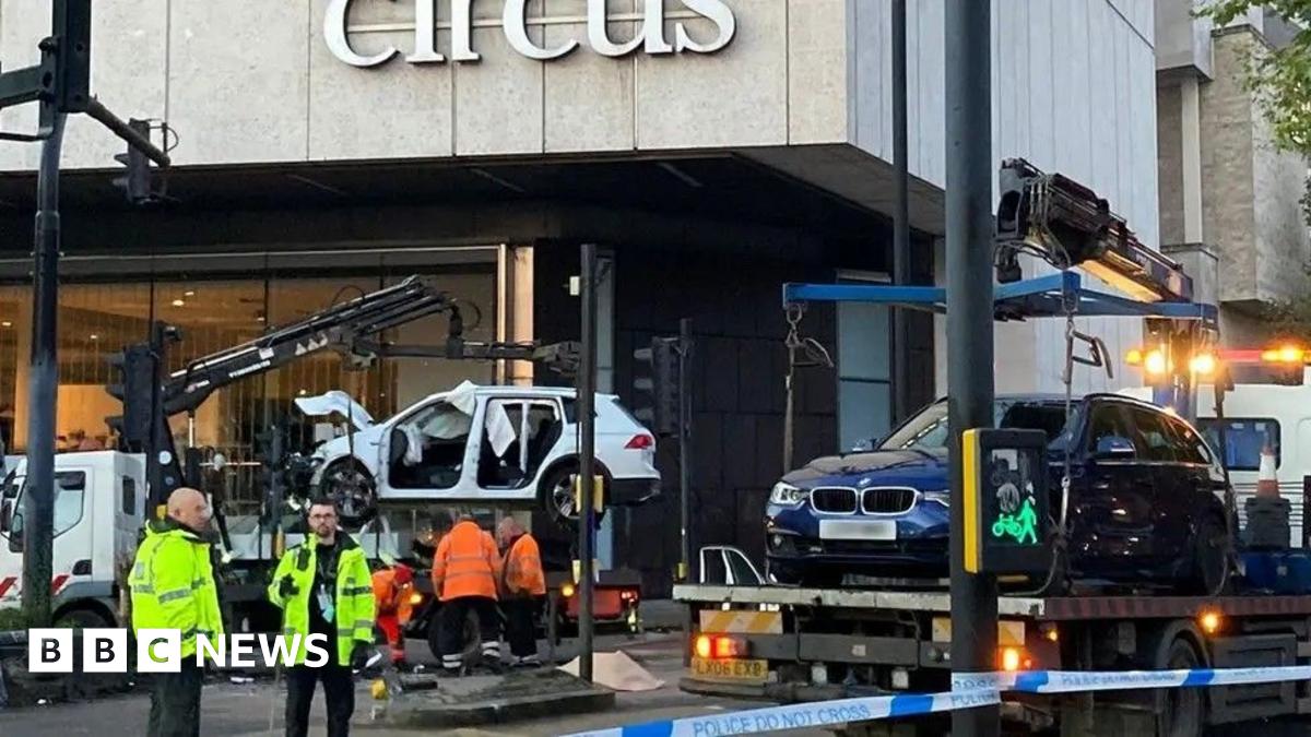 Members of a vehicle recovery team wearing yellow and orange hi-vis jackets. They are standing near the scene of the crash where a white Honda Jazz is being taken away on the bed of a crane truck. The blue BMW is visible on the right, also being removed from the scene.