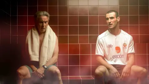 Aidan Monaghan Steve Coogan, playing Mick McCarthy wearing shorts in a sauna, with red tiles, next to Éanna Hardwicke, playing Mick McCarthy, who has his full Republic of Ireland kit on, despite the heat.