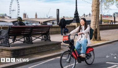 Woman rides a Santander bike on a London road with Big Ben and the London Eye in the background. She smiles and wears a white coat and jeans.