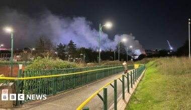 A footpath bordered by green metal railings with a yellow handrail. A pedestrian can been seen walking on the path. The path is lit up by overhead street lighting. A plume of smoke can be seen rising into the night sky.