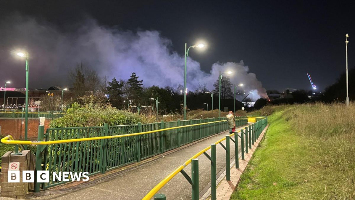 A footpath bordered by green metal railings with a yellow handrail. A pedestrian can been seen walking on the path. The path is lit up by overhead street lighting. A plume of smoke can be seen rising into the night sky.