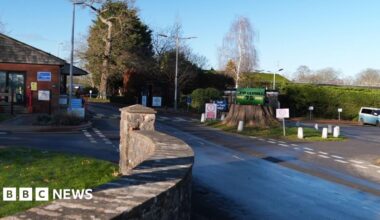 The road leading towards the entrance to HMP Leyhill on a sunny day. At the front of the image is a low stone wall. There is a large tree stump just right of the centre which has a green sign on it saying 'HMP Leyhill 75 1946 2021'. There are trees in the background, a small redbrick building to the left of the image and a blue and a white camper van parked to the right.