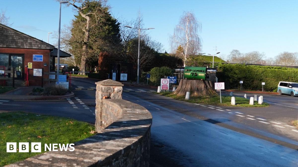 The road leading towards the entrance to HMP Leyhill on a sunny day. At the front of the image is a low stone wall. There is a large tree stump just right of the centre which has a green sign on it saying 'HMP Leyhill 75 1946 2021'. There are trees in the background, a small redbrick building to the left of the image and a blue and a white camper van parked to the right.