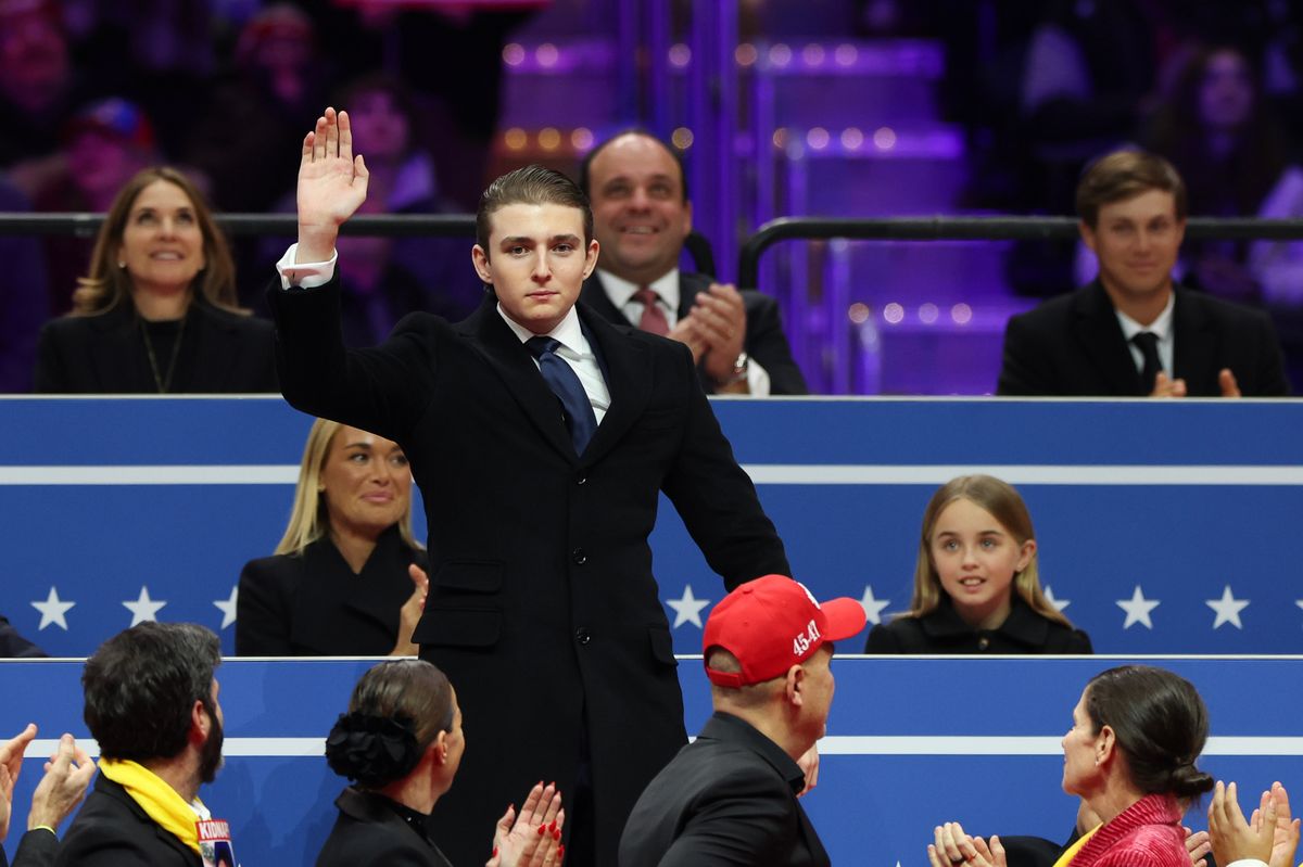 Barron Trump in a suit and navy tie waving to crowds as everyone else applauds him