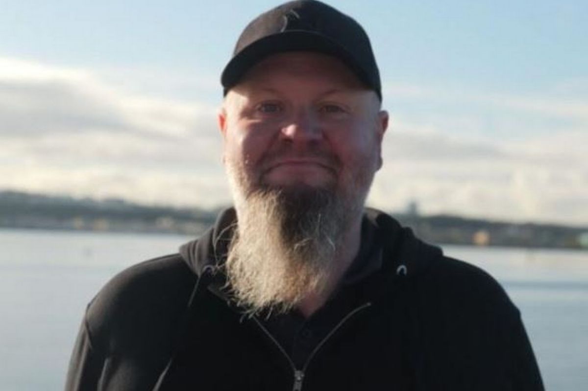 A man with a beard smiling at the camera while wearing a black baseball hat and a black hoodie