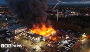 A fire is blazing from a building in an industrial estate. In there car park there are a large number of cars. One fire engine appears to be tackling the blaze