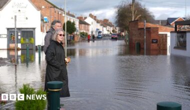 A man and a woman, both in winter coats, survey the flood waters in Station Road, Quorn, which is around ankle depth. Ripples can be seen in the water and the Monk restaurant partly submerged in the background.