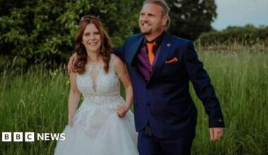 Kayleigh and Christopher Coll walking through a field of long grass and smiling - Mrs Coll is wearing a white wedding dress and Mr Coll is wearing a dark blue suit, purple waistcoat and orange tie.