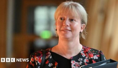 Shona Robison, with blonde hair and wearing a black top and patterned jacket, walks in the Scottish Parliament, carrying a black folder.