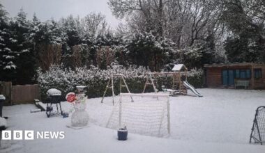 A "stop here santa" Christmas decoration covered in snow looks like a snowman in a garden. There is play equipment and everything is snowy.