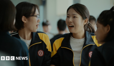 Four high school girls sit together in a cafeteria. They are wearing matching black-and-yellow athletic-school school jackets. Joo-in's friend, left, is turned slightly to the side wearing glasses, while Joo-in faces forward with her mouth open mid-expression