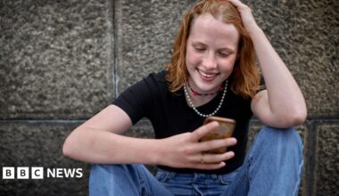 A girl looks at her phone, she has her hand on her head smiling and is wearning a black top and blue jeans.