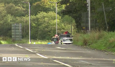 A road with trees either side and the top of some emergency service vehicles at the scene of a crash are just visible.