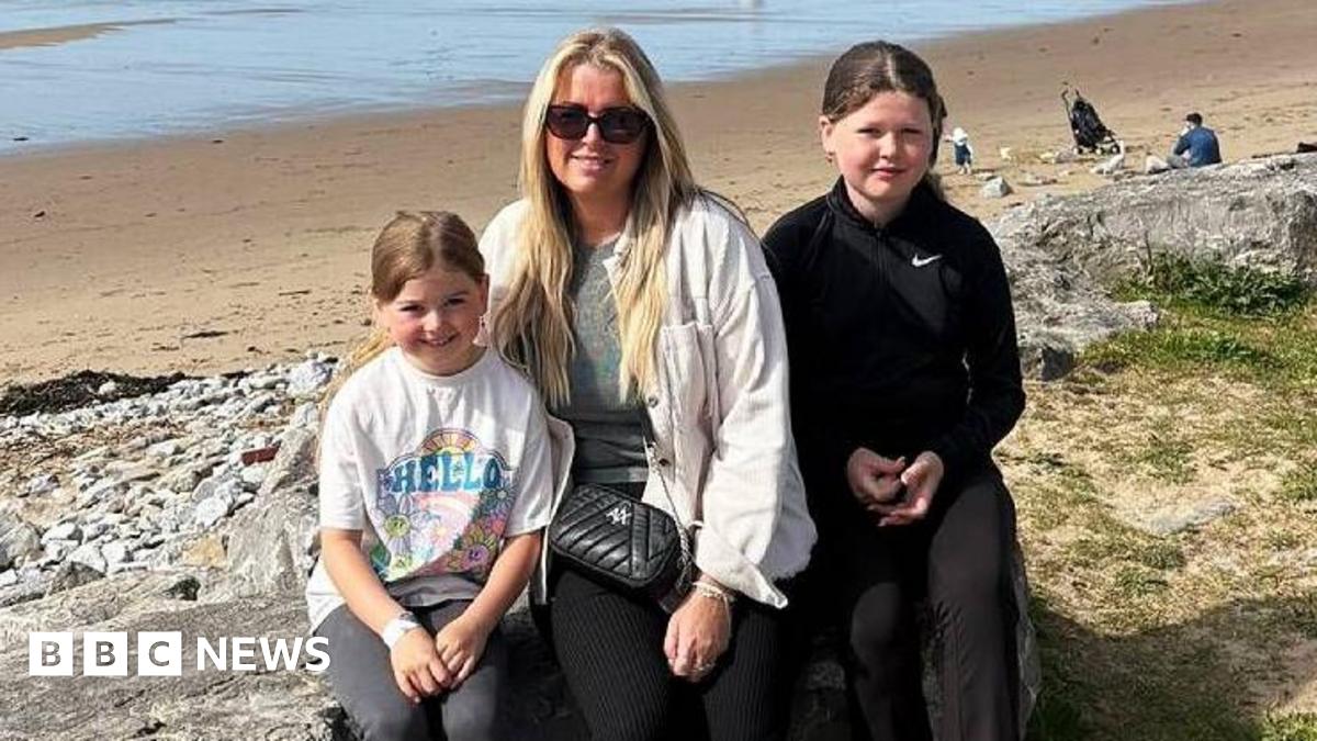 A young white girl wearing a t-shirt saying hello, her mother and her older sister sit on a rock at the edge of the beach. The mother, Lucy, has long blonde hair and wears black trousers, a plain tshirt and a white light jacket and black handbag over it. She is wearing sunglasses. Lola to her right is aged about 11, is wearing black trousers and sport top. all three are smiling at the camera. It's a sunny day. A few people sit on the beach behind them.