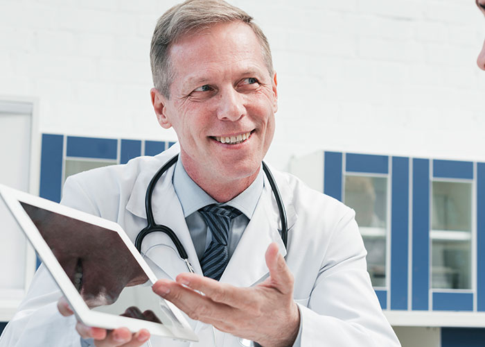 Smiling male doctor in white coat showing a tablet in medical office, illustrating unprofessional doctor-patient interactions.