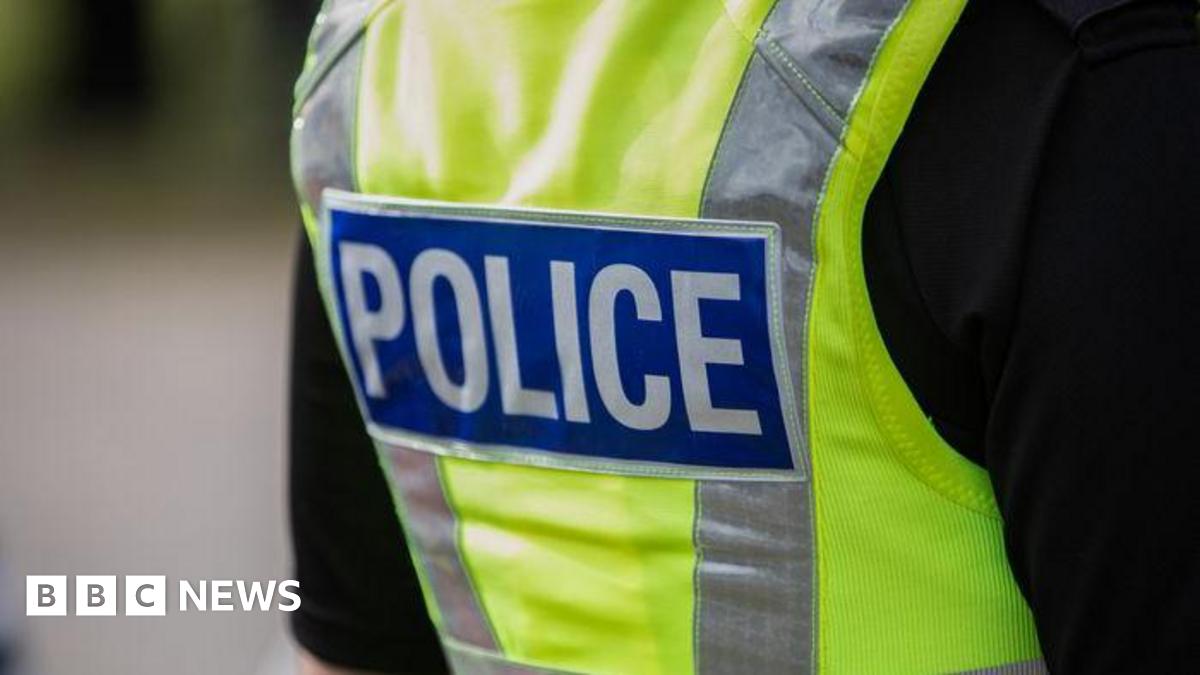 The upper back and shoulder area of someone wearing a high‑visibility yellow police vest. The vest features reflective strips and a large rectangular blue patch with the word “POLICE” written in bold white letters. The person is dressed in a black short‑sleeved shirt underneath the vest, and the background is softly out of focus, suggesting an outdoor setting.