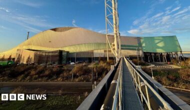 A large, modern building with a distinctive wave-like roof design, combining metallic and green-tinted panels. The structure appears industrial or possibly a sports or event venue, with a sleek, contemporary style. In the foreground, a metal walkway with railings extends toward the building, creating strong leading lines. To the right of the walkway, a tall, white structural tower rises vertically, adding to the industrial aesthetic. The surrounding area includes a road, landscaped greenery, and several parked vehicles near the entrance. The sky above is clear with scattered clouds, bathed in warm sunlight, giving the scene a bright and crisp appearance.