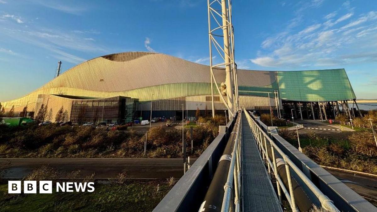 A large, modern building with a distinctive wave-like roof design, combining metallic and green-tinted panels. The structure appears industrial or possibly a sports or event venue, with a sleek, contemporary style. In the foreground, a metal walkway with railings extends toward the building, creating strong leading lines. To the right of the walkway, a tall, white structural tower rises vertically, adding to the industrial aesthetic. The surrounding area includes a road, landscaped greenery, and several parked vehicles near the entrance. The sky above is clear with scattered clouds, bathed in warm sunlight, giving the scene a bright and crisp appearance.