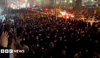 A view from a balcony at night of a street filled with people, stretching into the distance, marching towards the camera. The road is lit by street lights, car headlights and lights from buildings. To the left is a balcony where people are watching. A woman nearby is clapping.