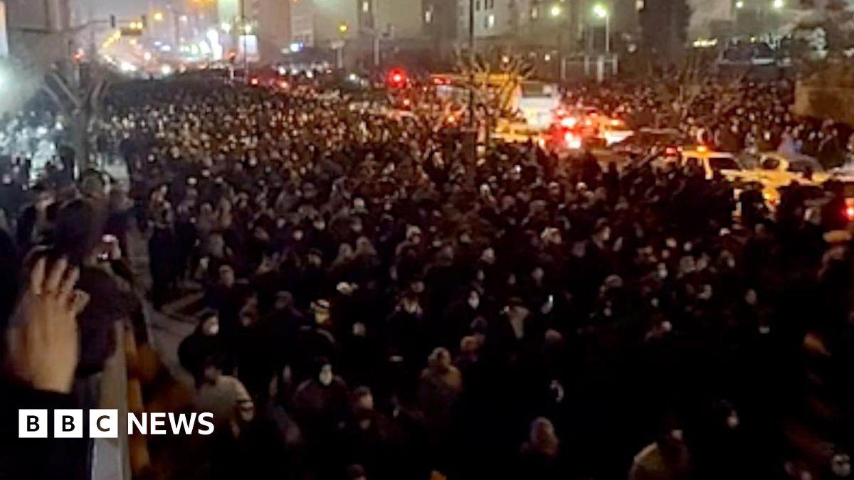 A view from a balcony at night of a street filled with people, stretching into the distance, marching towards the camera. The road is lit by street lights, car headlights and lights from buildings. To the left is a balcony where people are watching. A woman nearby is clapping.