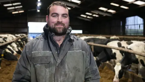 BBC Gethin Hughes, dairy farmer stands in his cattle shed wearing blue overalls