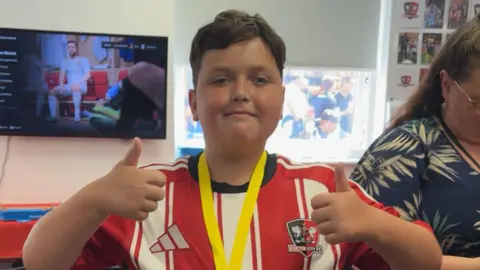 Family photo Oliver looking towards the camera with his thumbs up. He is smiling and is wearing a red and white striped shirt. There is a Exeter City FC badge on the right side of the shirt. Behind him is a TV and a woman stood to his right.