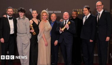 Doherty standing on a stage at the awards ceremony. She is holding a Golden Globe trophy in the left hand while speaking into a microphone positioned on a stand in front of her. She is wearing a sleeveless black dress with a fitted bodice and a decorative bow detail near the waist. A bracelet is visible on the right wrist, and earrings can also be seen. The background features a patterned wall with vertical panels and several spherical decorative elements.