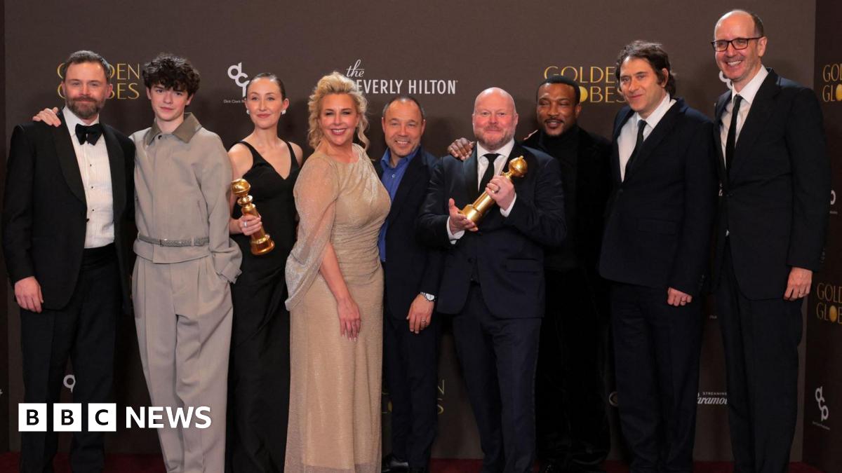 Doherty standing on a stage at the awards ceremony. She is holding a Golden Globe trophy in the left hand while speaking into a microphone positioned on a stand in front of her. She is wearing a sleeveless black dress with a fitted bodice and a decorative bow detail near the waist. A bracelet is visible on the right wrist, and earrings can also be seen. The background features a patterned wall with vertical panels and several spherical decorative elements.