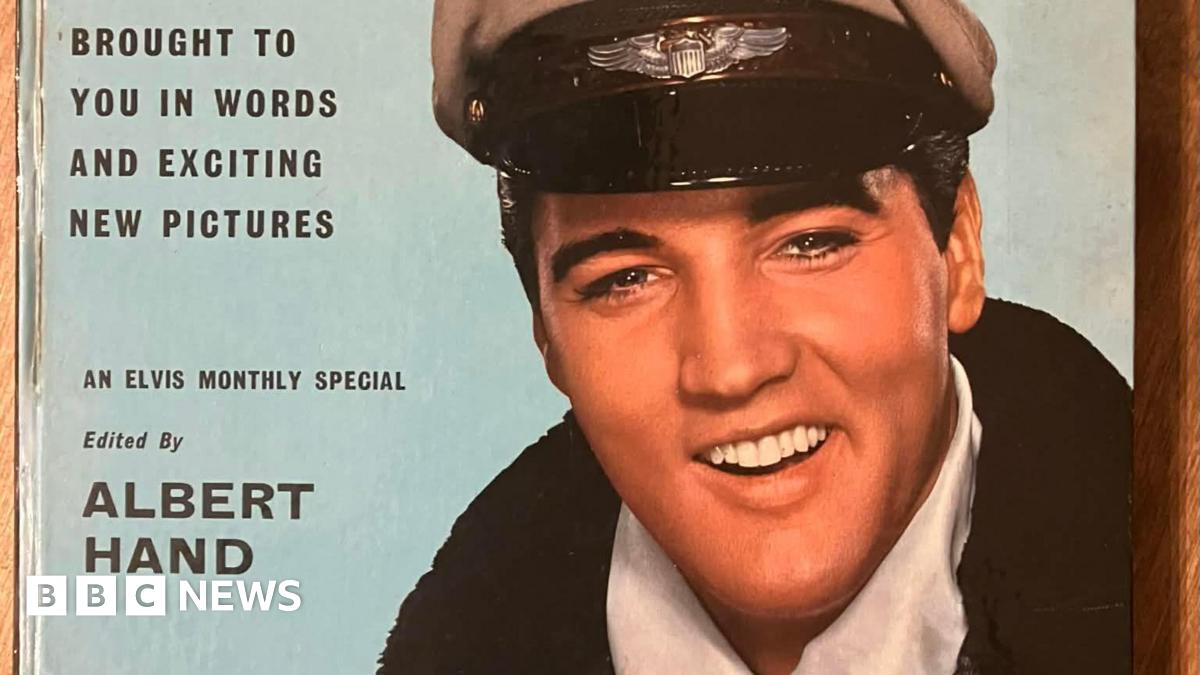 Part of a hardback magazine cover with a colour image of Elvis Presley wearing a military cap, white scarf and black coat. The cover of the magazine is light blue. In black lettering, its says, "...entertainer brought to you in words and exciting new pictures. An Elvis monthly special. Edited by Albert Hand."