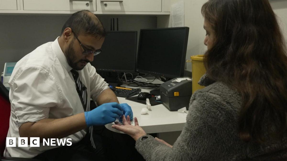 A man wearing a white shirt, tie and blue gloves takes a finger-prick blood sample from a woman seated across a desk, with computer monitors and medical equipment visible in the room.