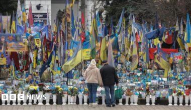 A woman and a man stand in front of a memorial for fallen soldiers in Kyiv, Ukraine, with flags and photographs.