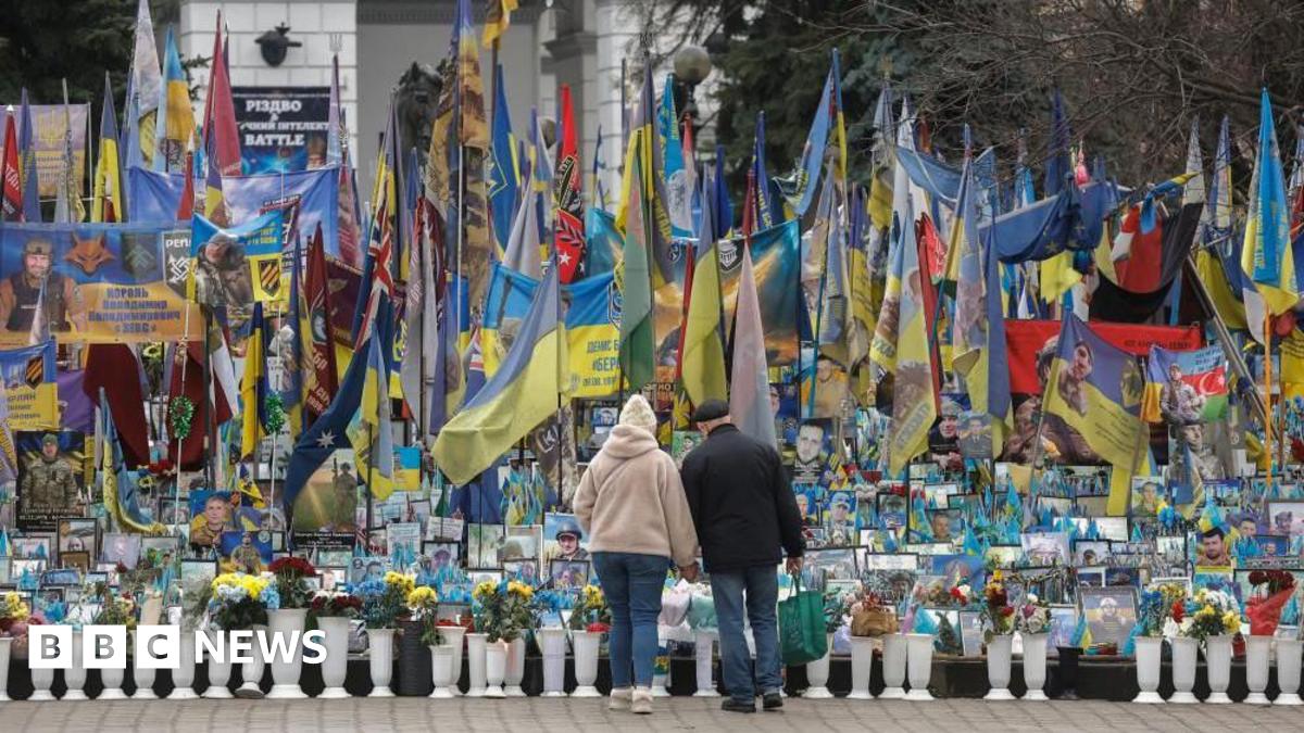 A woman and a man stand in front of a memorial for fallen soldiers in Kyiv, Ukraine, with flags and photographs.