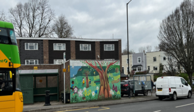 A view of a bus stop on a grey day: red brick building behind with windows and a flat roof. The bus stops have a bright mural painted next to them, of a cheery green park with flowers and children playing. On the left, the front of a yellow and green bus starts to come into view