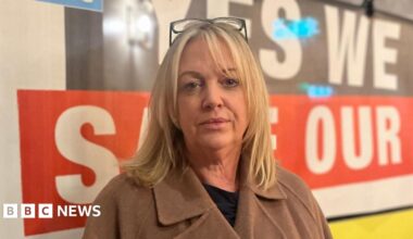 Kate is looking directly at the camera. She has shoulder-length blond hair and has a pair of reading glasses on her head. She is wearing a black t-shirt and brown coat.