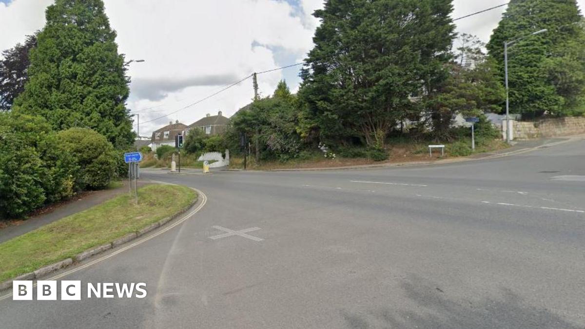 A crossroads junction in a residential area. There is a street name sign at one corner and there is a green verge and tress obscuring houses.