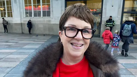 Karen Jones is outside Cardiff Central station and is wearing glasses a brown winter coat and a red Christmas sweater