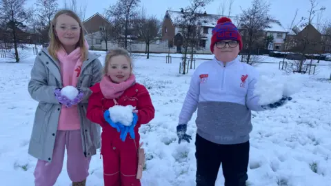 Three children standing side by side with snowballs in their hands. They are all wearing gloves as they stand in a garden covered with thick snow. The two girls have long blonde hair and the boy is wearing a hat with glasses.