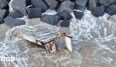 Millie Bobby Brown and David Harbour in character as Eleven and Jim Hopper. Eleven wears red shorts over grey leggings and a grey jumper. Jim Hopper wears a black leather jacket, a black beanie hat and dark trousers.They are speaking to director Frank Darabont on the set of Stranger Things. There is a vintage advertisement posted in the background with debrief on the ground.