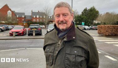 A man with grey hair, wearing a dark green jacket, smiles at the camera with hsi arms behind his back. A car park is behind him.