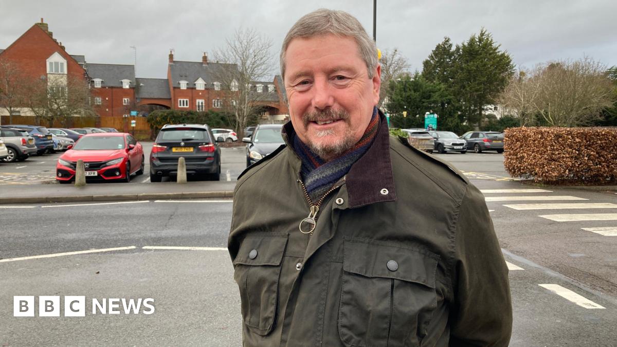 A man with grey hair, wearing a dark green jacket, smiles at the camera with hsi arms behind his back. A car park is behind him.