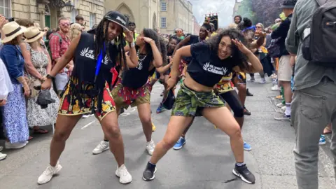 A troop of street dancers in the parade at the 2023 St Pauls carnival, they are wearing black t shirts with the St Pauls Carnival logo and wear colourful skirts, they appear to be striking a salute pose and as part of their dance
