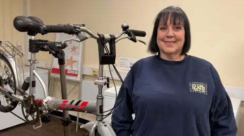 BBC News  A lady wearing a navy jumper stands in front of a bike which is being held up by a metal stand. She has black shoulder-length hair with a fringe and is smiling at the camera. The silver bike has a black seat and handlebars.