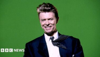 David Bowie is pictured in a navy blue suit, with a white shift and navy and white tie, in front of a bright green background. He's smiling, and the tie is flipped back over one shoulder