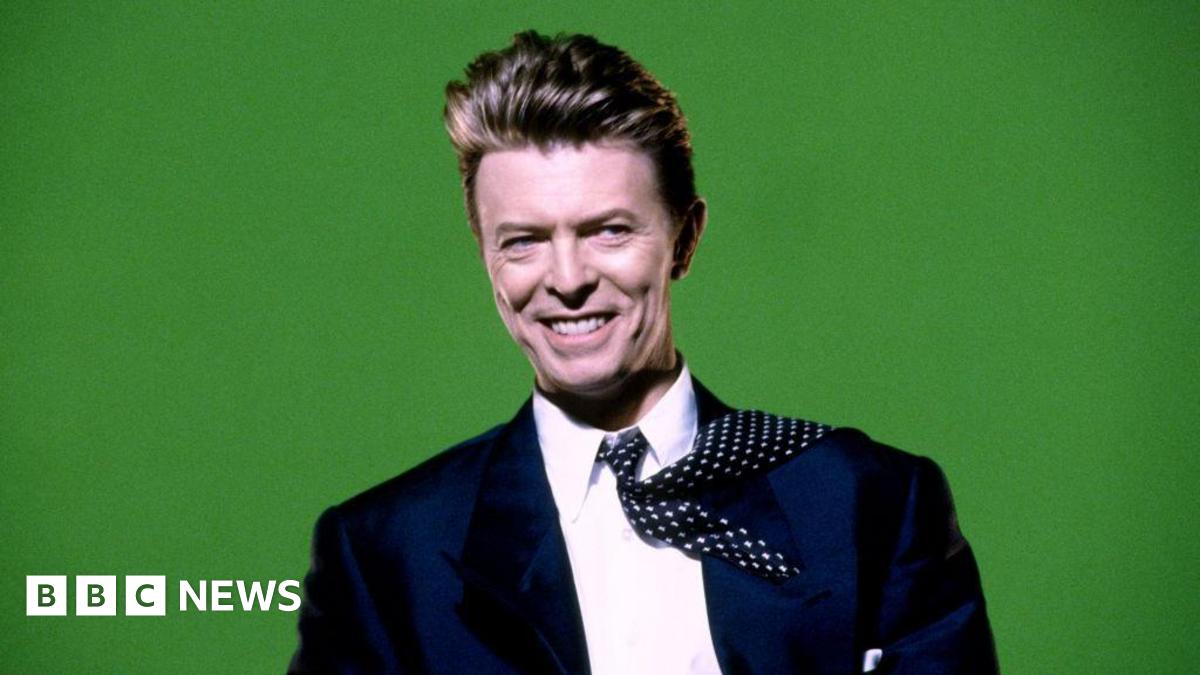 David Bowie is pictured in a navy blue suit, with a white shift and navy and white tie, in front of a bright green background. He's smiling, and the tie is flipped back over one shoulder
