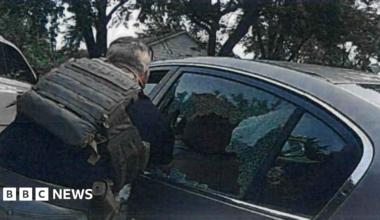 Person wearing tactical vest leans toward a car with shattered rear window, broken glass visible, trees and houses in background.