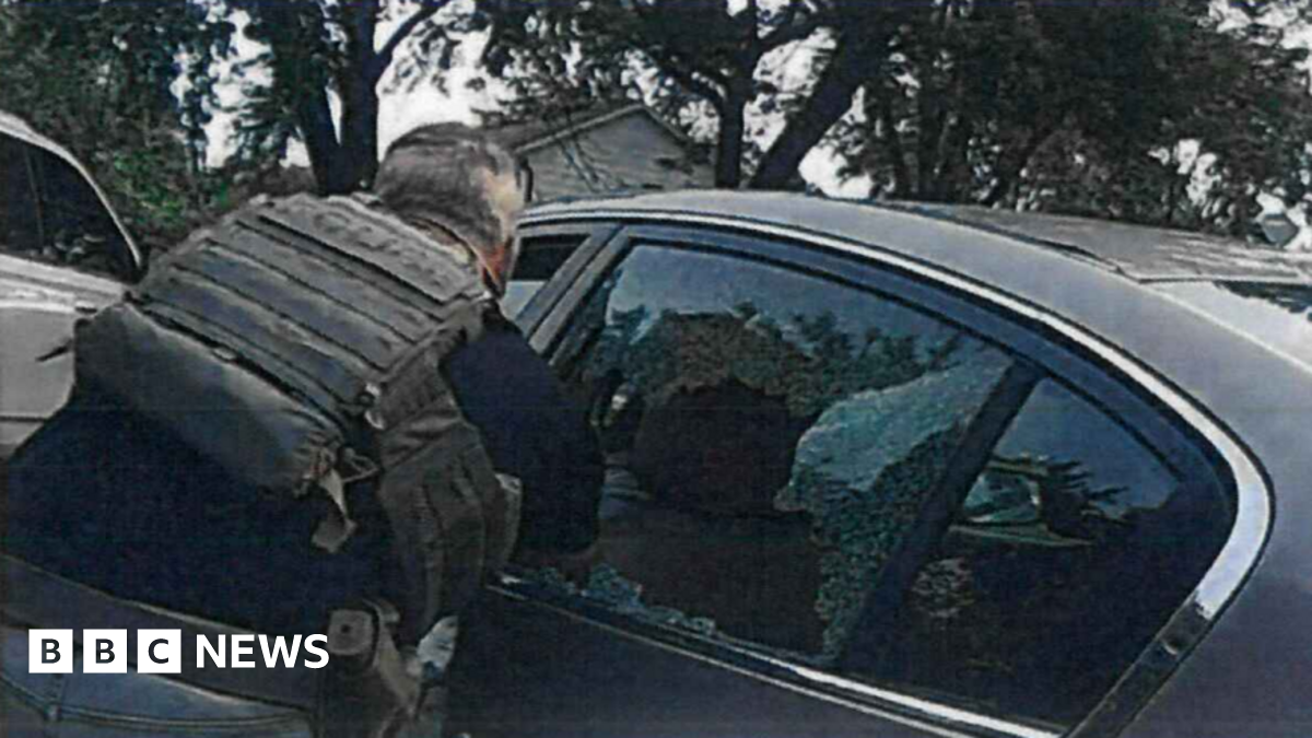 Person wearing tactical vest leans toward a car with shattered rear window, broken glass visible, trees and houses in background.