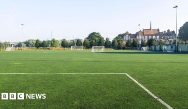 The image shows an empty leisure field in a residential district, with a view of the city in the distance. The sky is cloudless and light blue. The white lines of a football pitch can be seen in the foreground of the image.